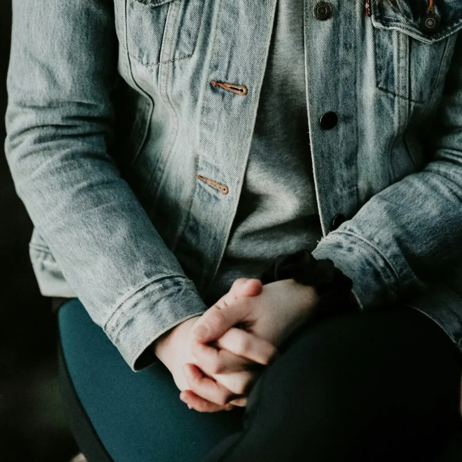 A person sitting in a denim jacket, hands clasped on their knees