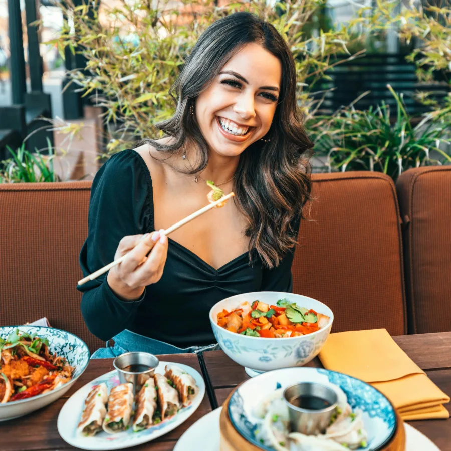 A smiling young woman at a restaurant, chopsticks in hand, in front of several dishes
